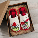 A pair of white sherpa slip-on slippers with red and black plaid trim, featuring Grinch and Sally the Christmas cartoon character graphics, presented in a brown gift box filled with paper straw on a light wood floor.