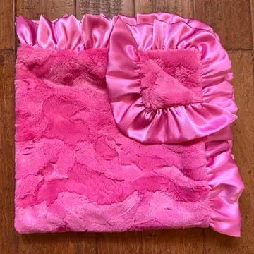 A folded pink blanket with a textured fuzzy front and a shiny satin ruffled corner, placed on a wooden floor.