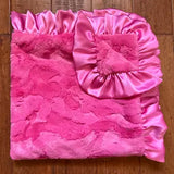 A folded pink blanket with a textured fuzzy front and a shiny satin ruffled corner, placed on a wooden floor.