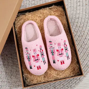 A pair of pink fuzzy slipper shoes with embroidered nutcracker figures, presented inside a brown gift box filled with shredded paper on a gray textured rug.