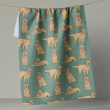 A kitchen towel with a repeating pattern of golden retriever dogs wearing pink bandanas on a muted teal background, hanging from a string against a neutral gray wall.
