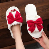 A pair of white fuzzy indoor slippers with large red bow details on the top, shown with one being worn on a foot and the other held by a hand against a dark wood background.