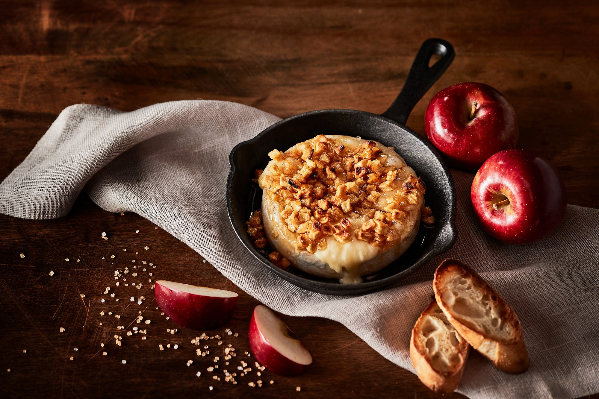 Small skillet with baked dish on a wooden surface with apples and bread slices.