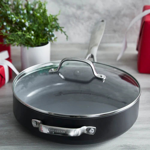 Black frying pan with glass lid on a light wooden surface with decorative items in the background.