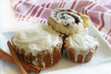 Three cinnamon rolls with cream cheese frosting on a white plate with a red and white striped towel in the background.