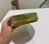 A hand holding a roll of decorative ribbon with a green center and textured gold edges, photographed against a white tabletop background.
