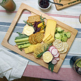 A wooden charcuterie board filled with assorted cheeses, crackers, sliced cucumbers, radishes, celery, and rosemary, arranged on a striped tablecloth in a real dining setting.