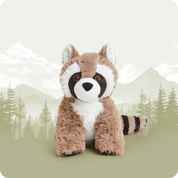 A plush raccoon toy centered against a soft, stylized forest and mountain backdrop. The raccoon has brown and white fur with black facial markings and a black-and-brown striped tail.