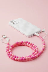 A pink beaded phone strap connecting a white smartphone to a matching white charging cable, arranged on a solid light pink background.