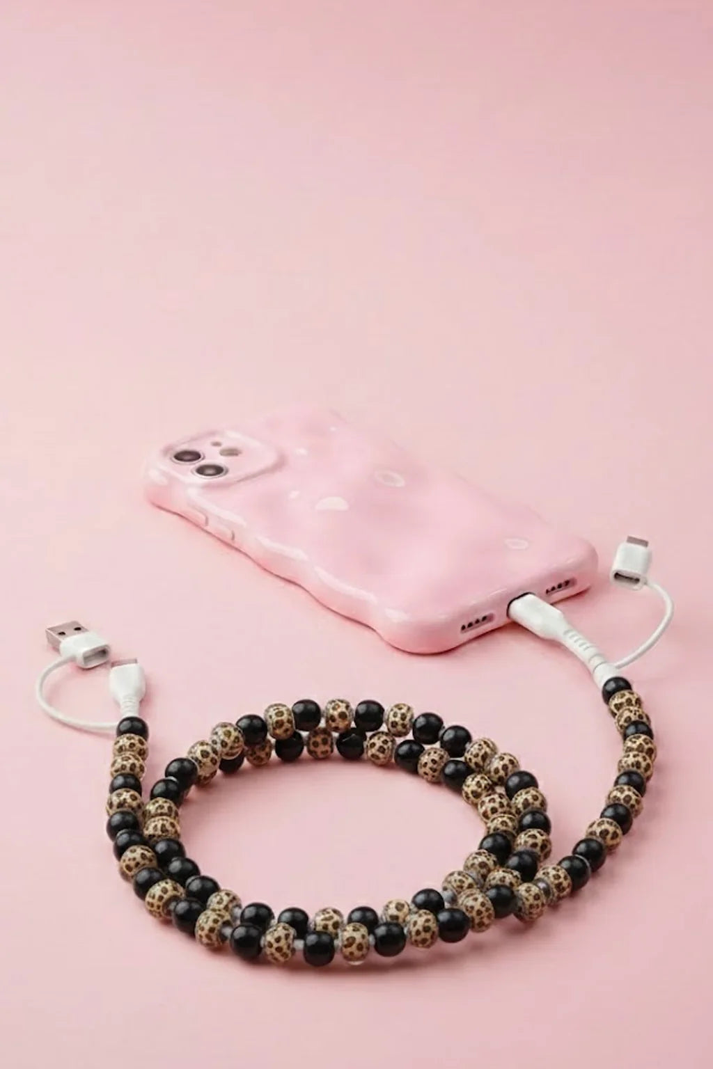 A pink phone case with a white charging cable attached, styled with a beaded phone strap made of black and leopard-print beads on a solid pink background.