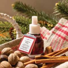 A bottle of Christmas room fragrance labeled 'Fragrance Magic' by Candle Matters, styled with cinnamon sticks, walnuts, pine branches, and a red-and-white checkered cloth.