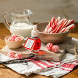 A small bottle of red peppermint liquid food coloring with a white cap and a red-and-white label, styled on a wooden board with candy canes, peppermint candies, vanilla beans, a pitcher of milk, and a striped cloth on a wooden table.