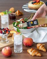 Table setting with peaches, croissants, salads, and a hand holding a Ouelleve peach product.