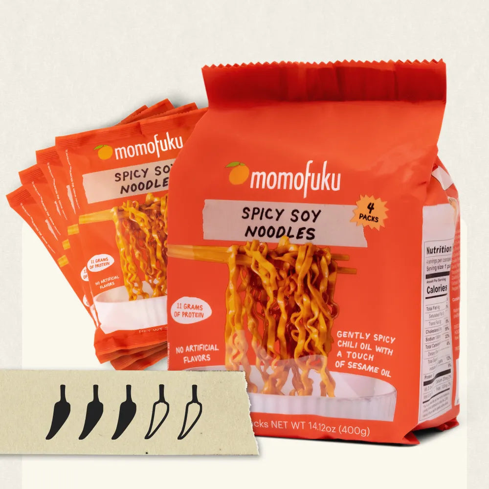 A package of momofuku Spicy Soy Noodles in a bright orange bag, with three additional bags fanned behind it on a light background. A small card with five pepper-shaped icons appears at the bottom left.