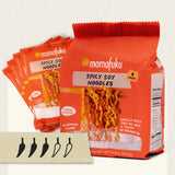 A package of momofuku Spicy Soy Noodles in a bright orange bag, with three additional bags fanned behind it on a light background. A small card with five pepper-shaped icons appears at the bottom left.