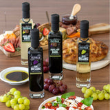 Four bottles of Olivelle balsamic vinegar on a table with food and grapes.