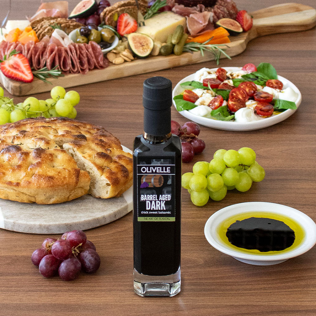 Olivelle dark olive oil bottle on a wooden table with bread, grapes, and salad.