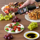 Person pouring olive oil over a salad with a charcuterie board and grapes on a wooden table.