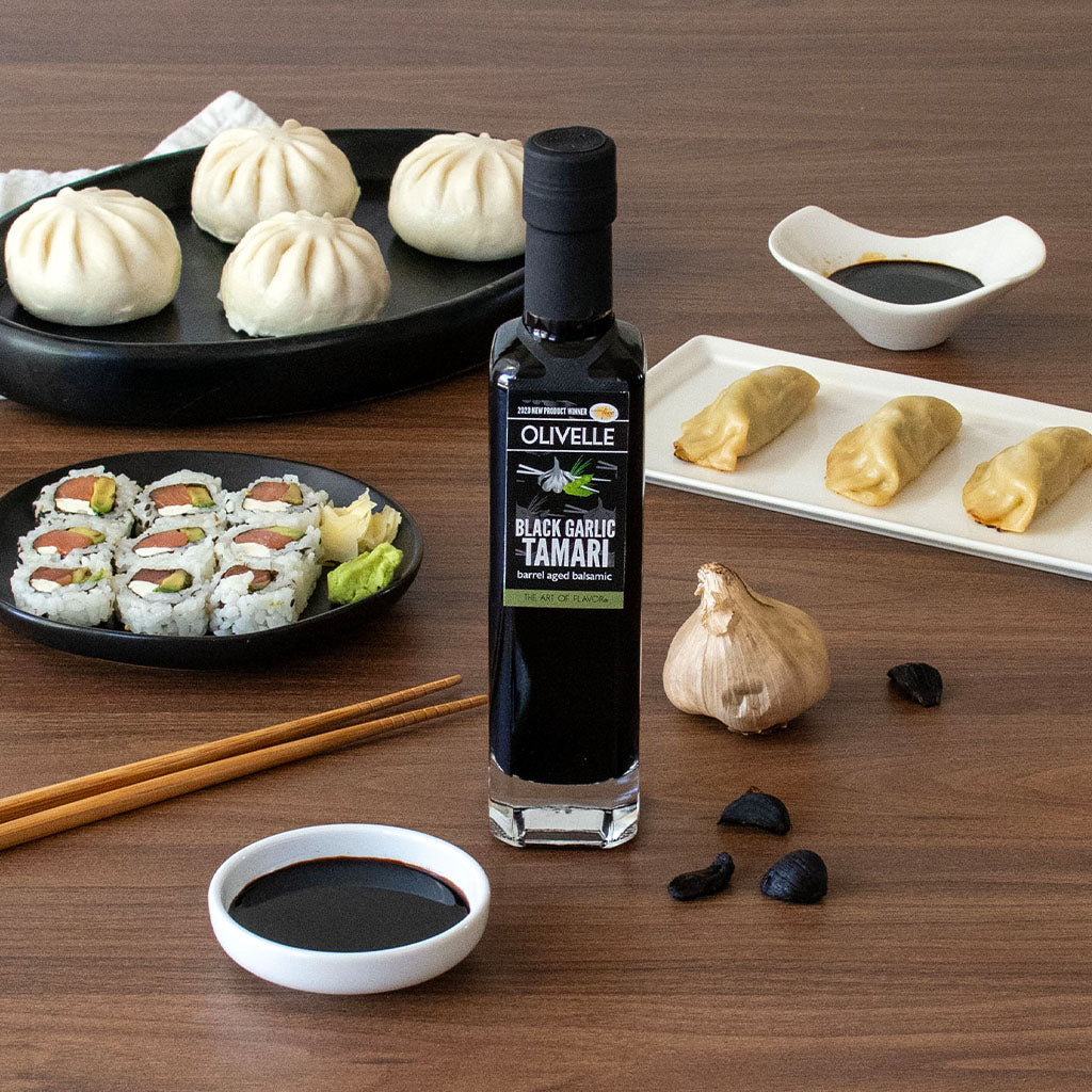 Olivelle Black Garlic Tamari bottle on a wooden table with sushi, dumplings, and a garlic bulb.