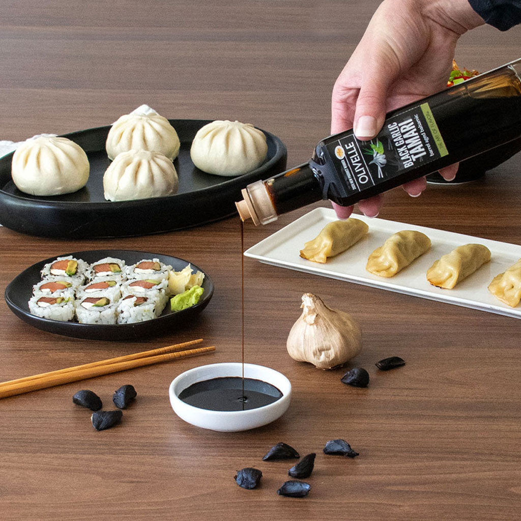 Person pouring olive oil over food on a wooden table with various dishes including sushi and dumplings.