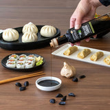 Person pouring olive oil over food on a wooden table with various dishes including sushi and dumplings.
