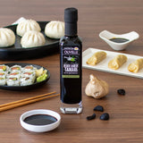 Olivelle Black Garlic Tamari bottle on a wooden table with sushi, dumplings, and garlic.