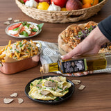 Person pouring olive oil over a dish on a wooden table with various food items.