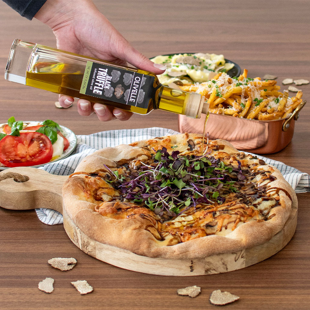 Person pouring olive oil over a pizza with salad and fries on a wooden table