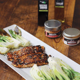 Plated dish of grilled salmon with sides on a wooden table, featuring sriracha and shiitake sauces.