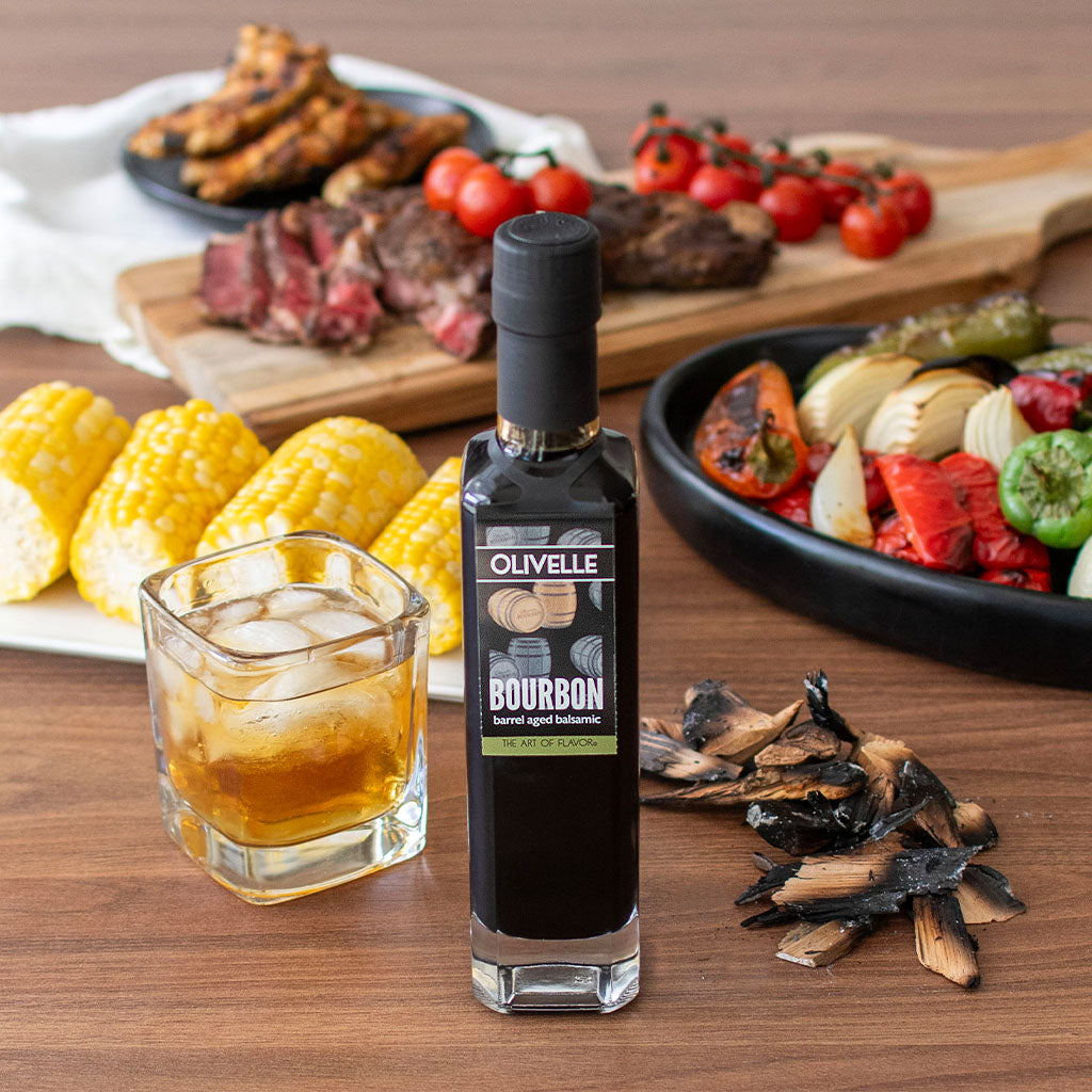 Bottle of Olivelle bourbon-aged balsamic vinegar on a wooden table with food and drinks in the background.