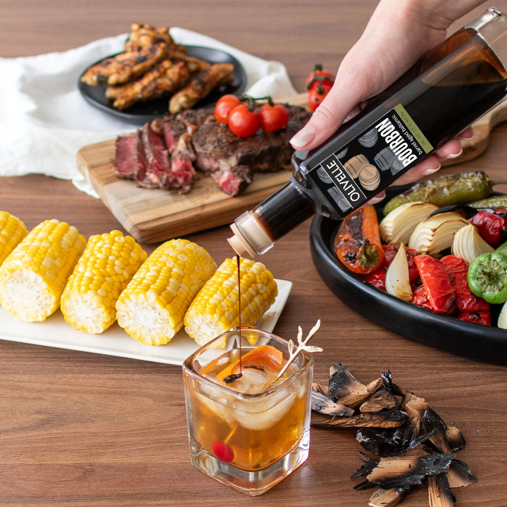 Person pouring a bottle of sauce into a glass on a table with food including corn, steak, and vegetables.