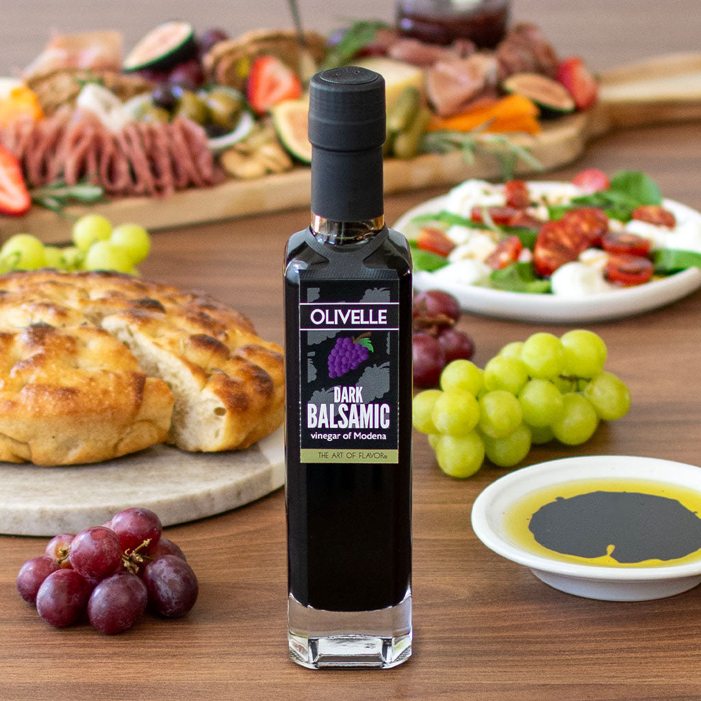 Olivelle dark balsamic vinegar bottle on a wooden table with food items in the background