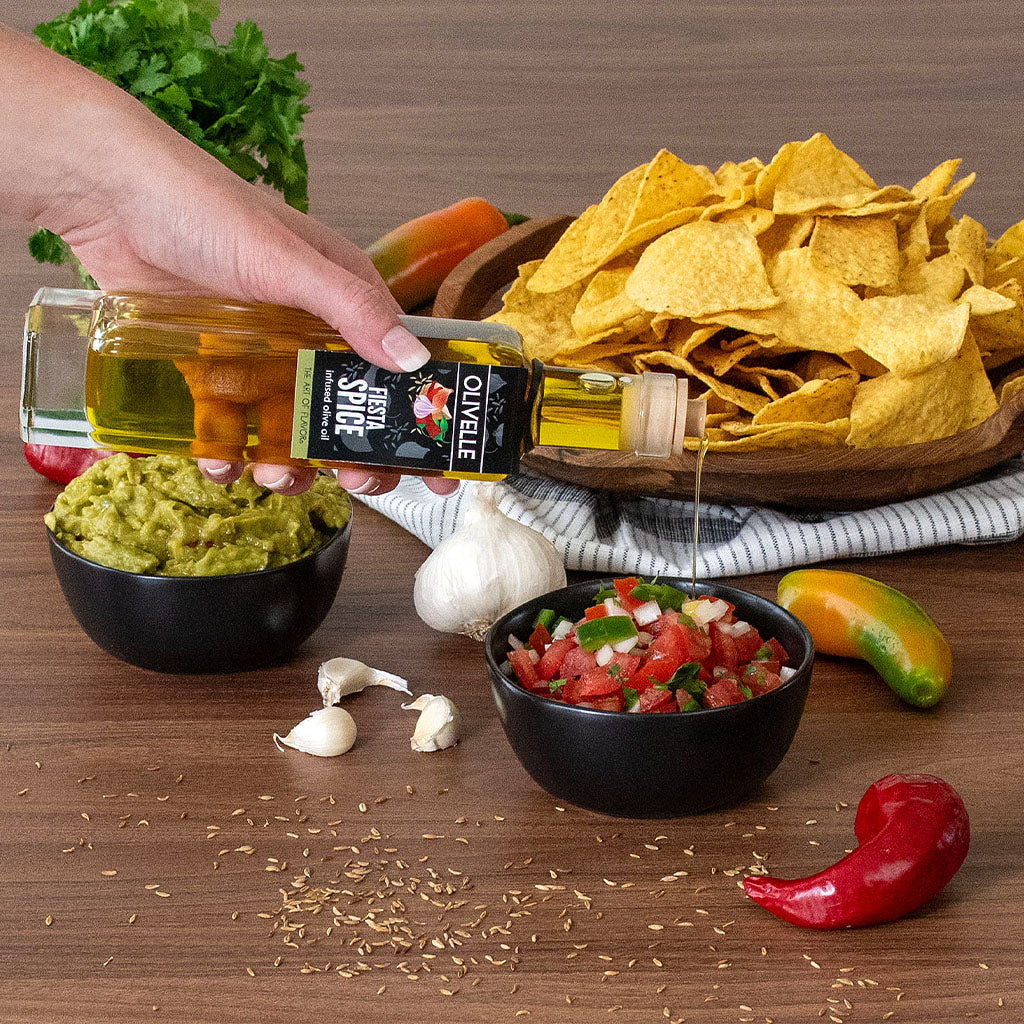 Hand pouring olive oil over guacamole with a bowl of chips, salsa, and fresh ingredients on a wooden surface.