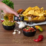 Hand pouring olive oil over guacamole with a bowl of chips, salsa, and fresh ingredients on a wooden surface.