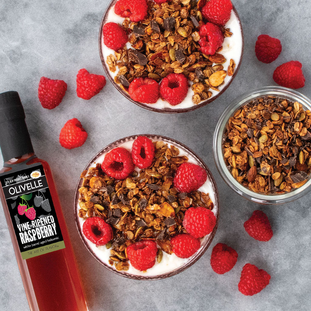 Two bowls of yogurt with granola and raspberries, a bottle of Olivelle raspberry vinegar, and additional raspberries on a gray surface.
