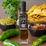 Bottle of Olivelle Green Chile olive oil with ingredients like chips, guacamole, and salsa on a wooden surface.