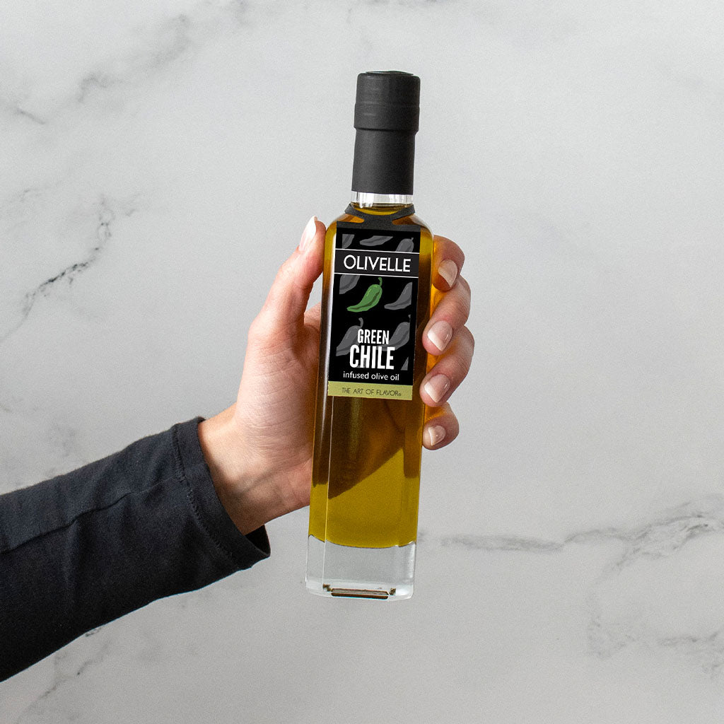 Hand holding a bottle of Olivelle Green Chile olive oil against a marble background