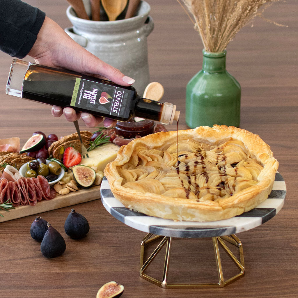 Person pouring fig syrup over a pie on a wooden table with figs and cheese platter.