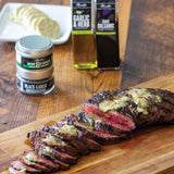 Sliced grilled steak on a wooden board with garlic and herb seasonings in the background.