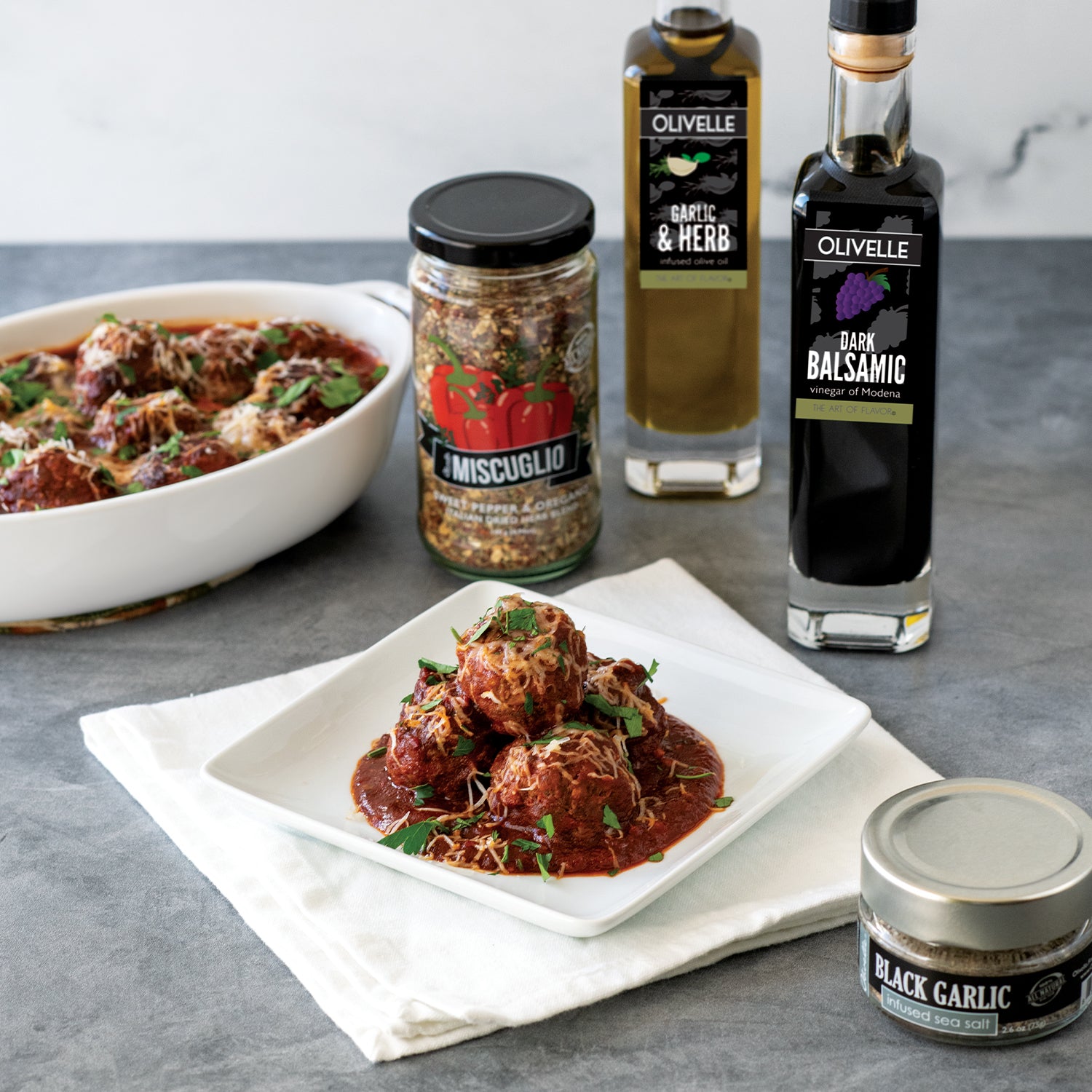 Plated dish of meatballs with red sauce, surrounded by Olvelle products on a gray surface.