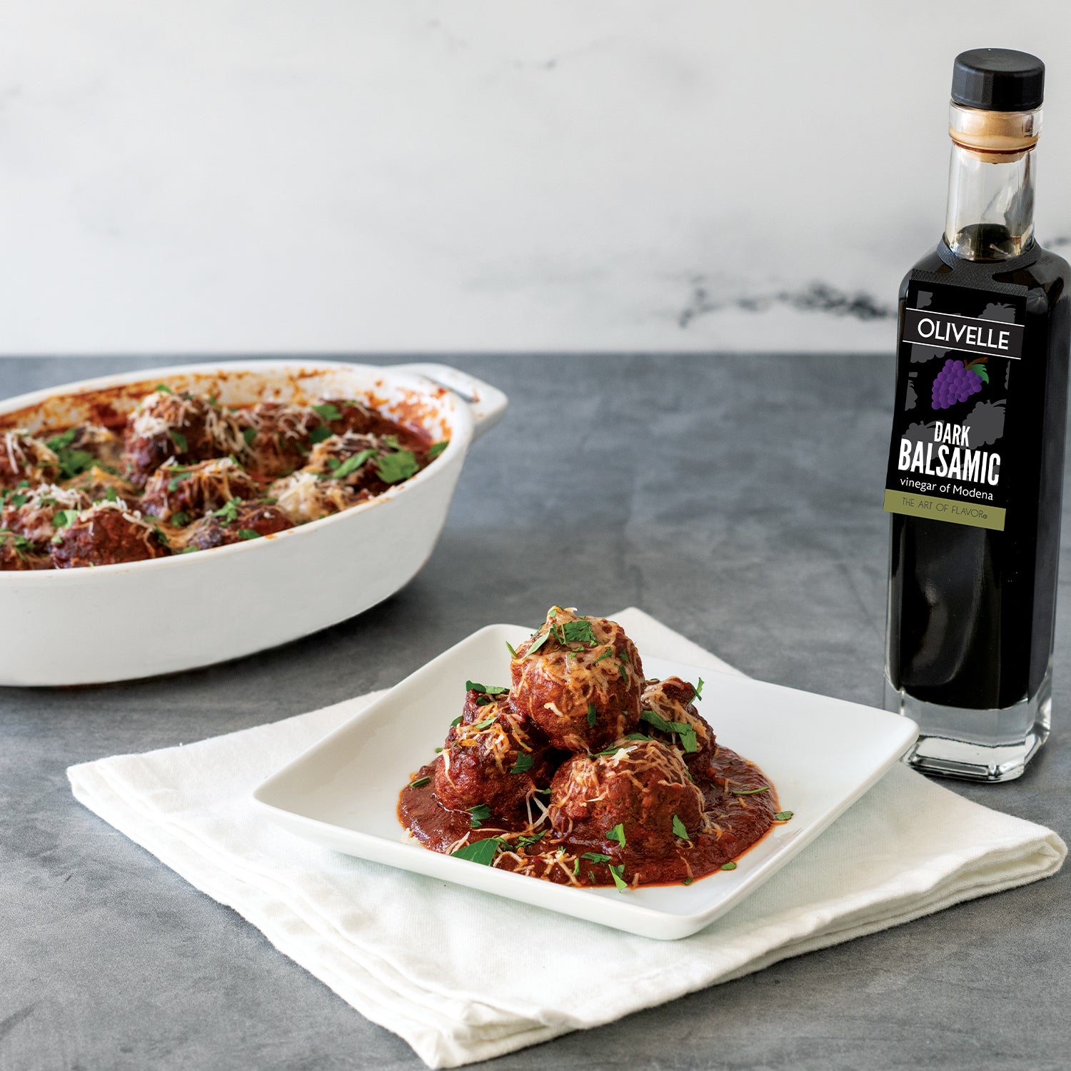 Bowl of meatballs with a bottle of Olivelle dark balsamic vinegar on a gray surface.