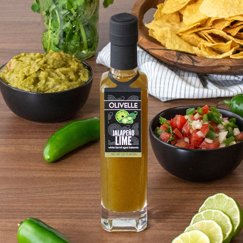 Bottle of Olivelle Jalapeno Lime dressing on a wooden table with guacamole, chips, salsa, and limes.
