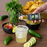 Person pouring Olivelle Jalapeño Lime sauce into a bowl of guacamole with a margarita, jalapeños, limes, and tortilla chips on a wooden table.