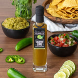 Olivelle Jalapeno Lime oil bottle with guacamole, chips, salsa, and limes on a wooden table.