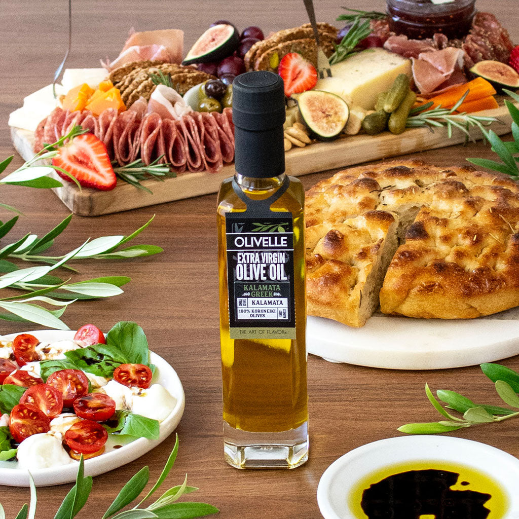 Bottle of olive oil on a wooden table with appetizers including bread, salad, and cured meats.