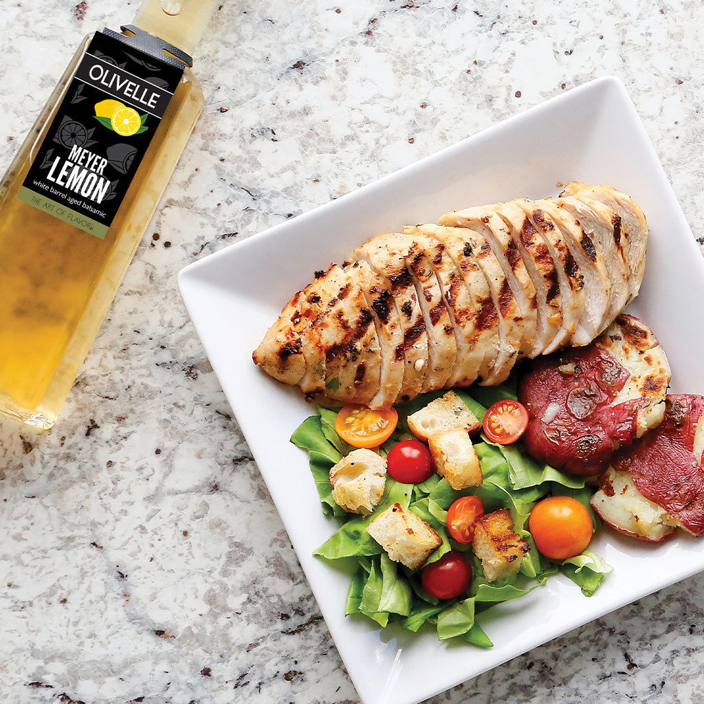 Plated dish of sliced grilled chicken breast with salad on a marble surface, featuring Olivelle Meyer Lemon olive oil.