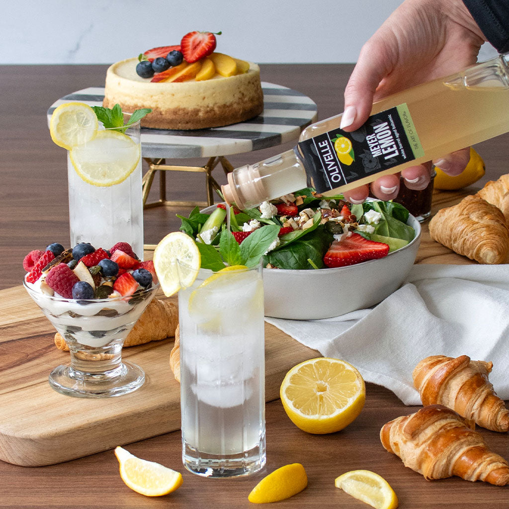 Person pouring lemon juice over a salad with a dessert, fruit, and pastries on a wooden table.