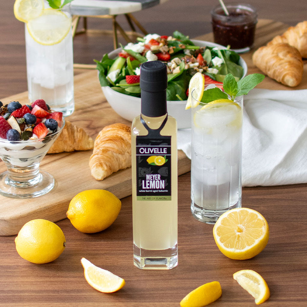 Olivelle Meyer Lemon bottle on a table with lemons, a salad, and other snacks.
