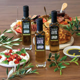 Three bottles of Olivelle olive oil on a wooden table with food in the background