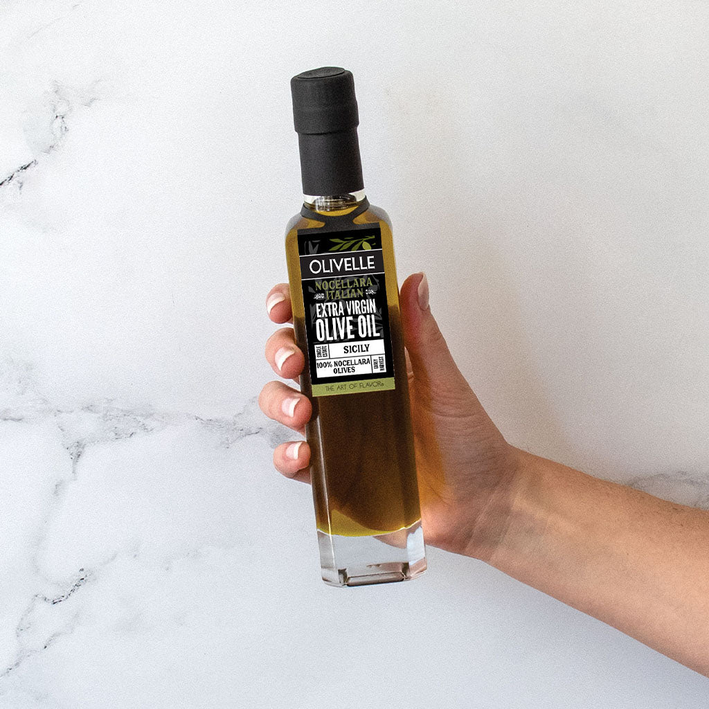 Hand holding a bottle of Olivelle olive oil against a white marble background